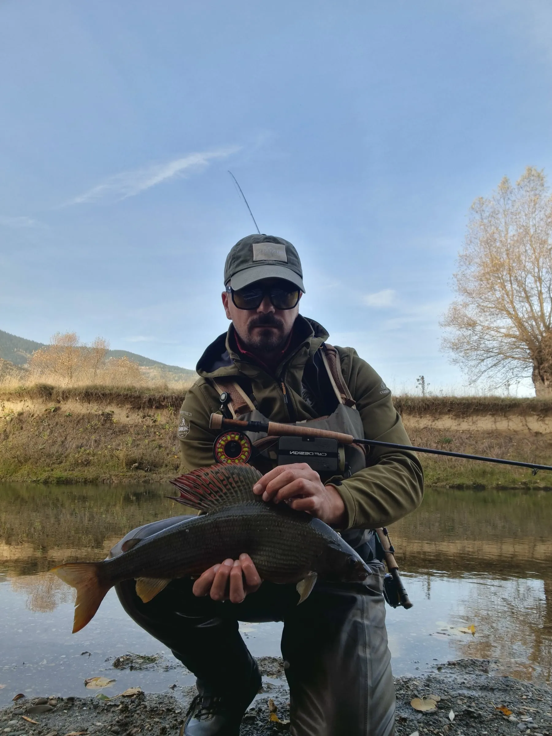 An angler crouching on a gravel bank of the Ljuča holding a large dark grayling with its fin spread, a willow and hills behind.