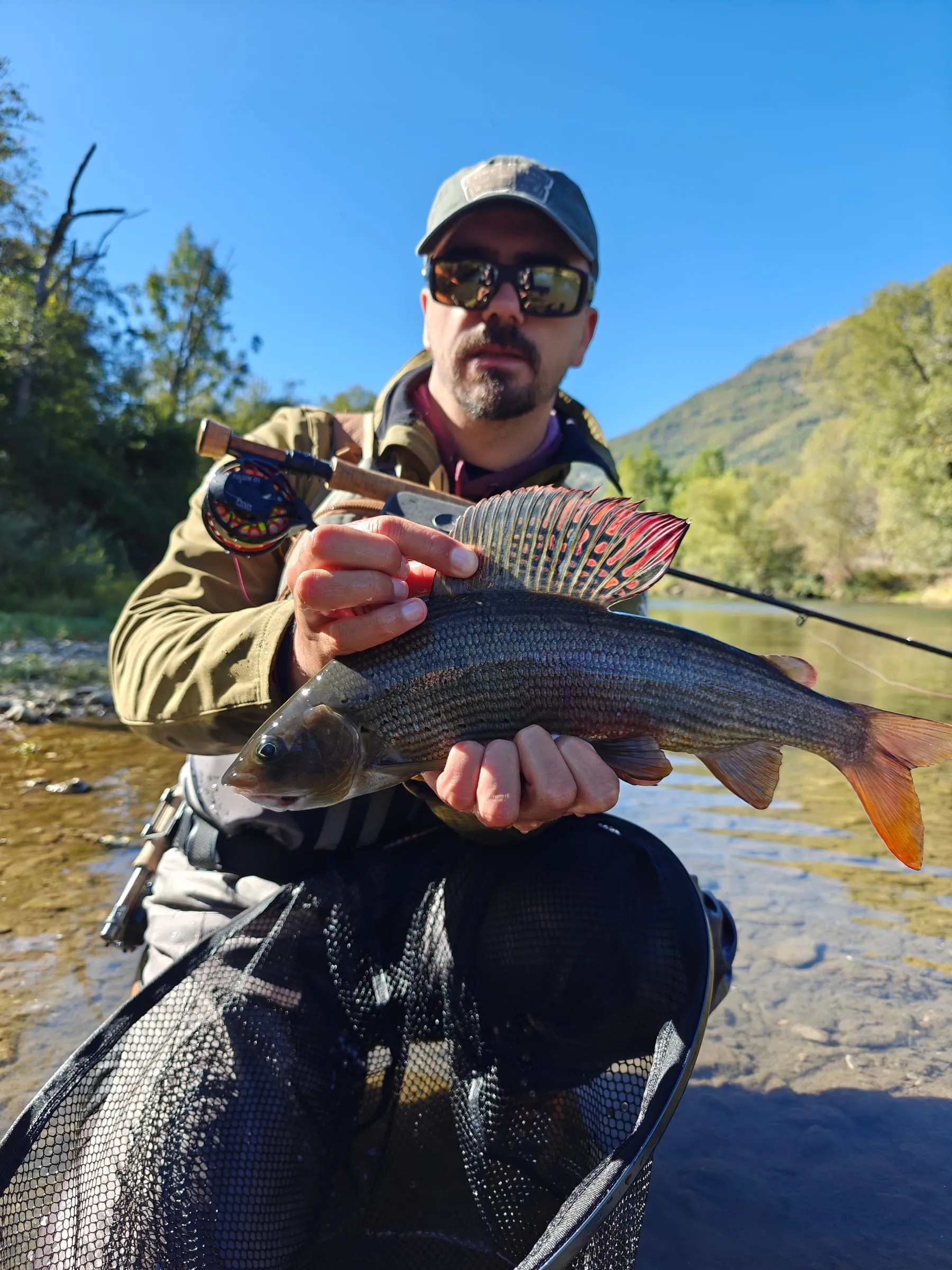 An angler holding a grayling with its red-spotted dorsal fin raised, the river and a green hillside behind.