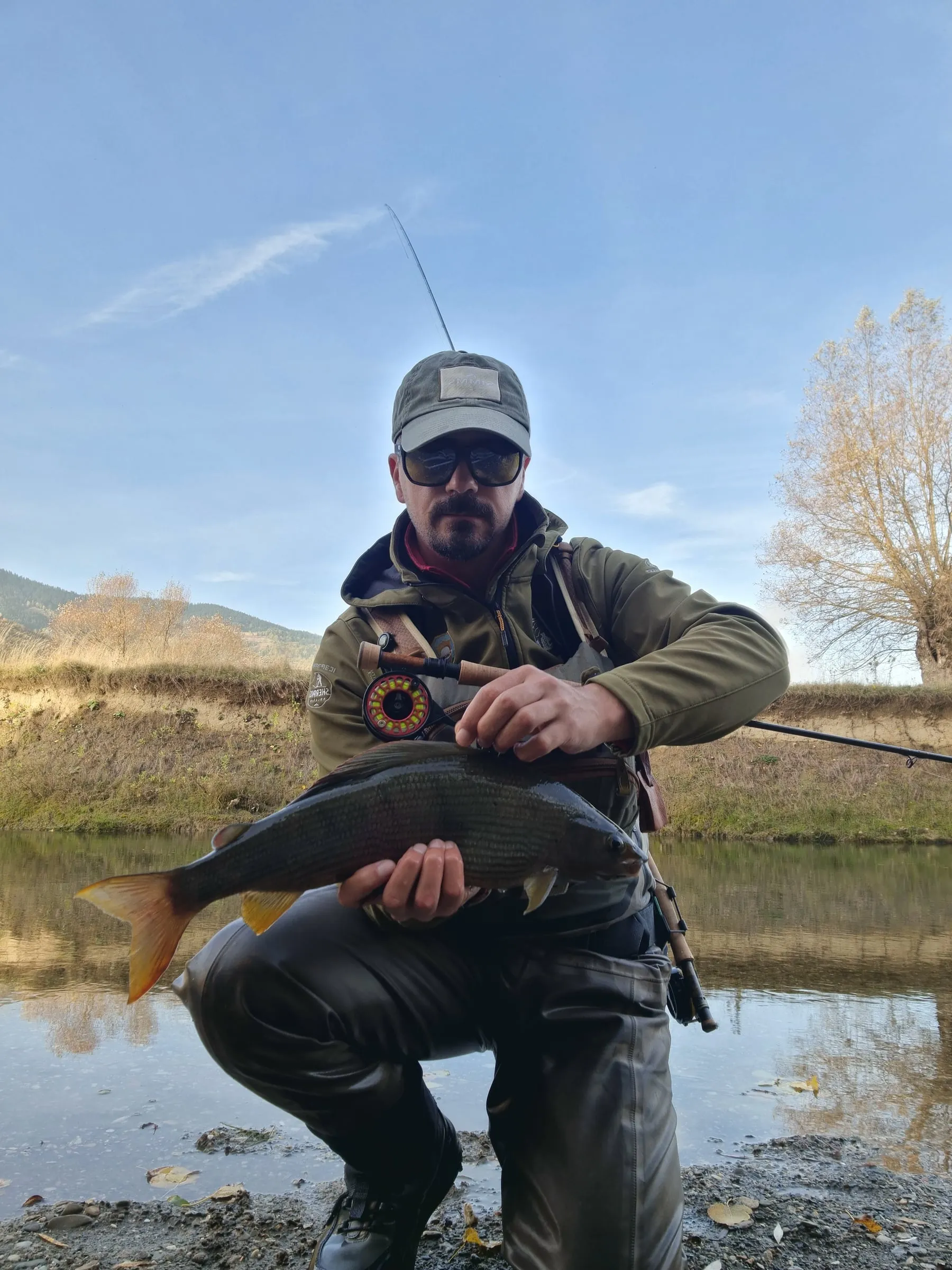 An angler crouching by the Ljuča holding a large grayling, a rod and reel at his chest, an autumn tree behind.