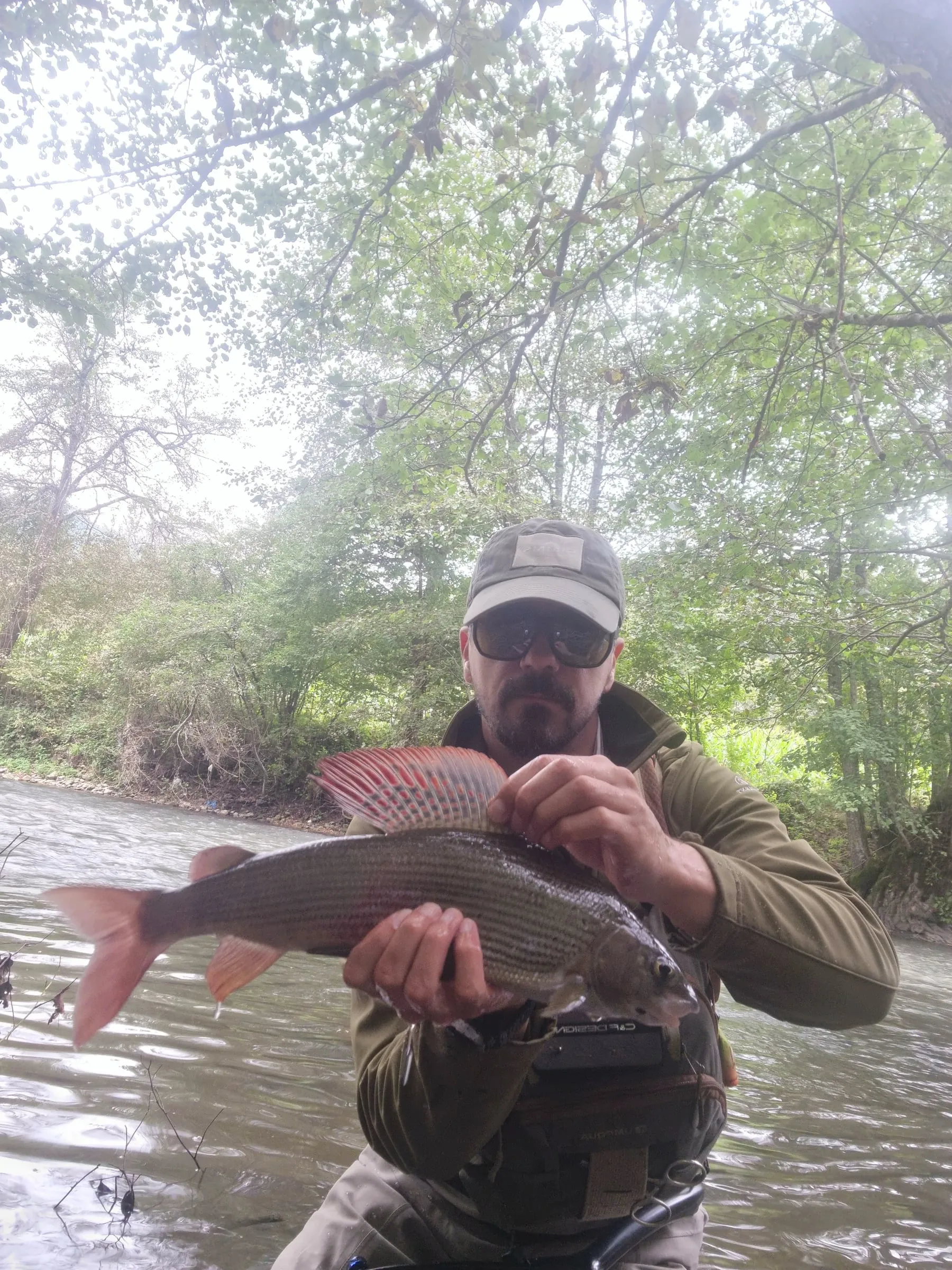 An angler in a cap and sunglasses holding a grayling with its tall dorsal fin raised, standing in the river under trees.