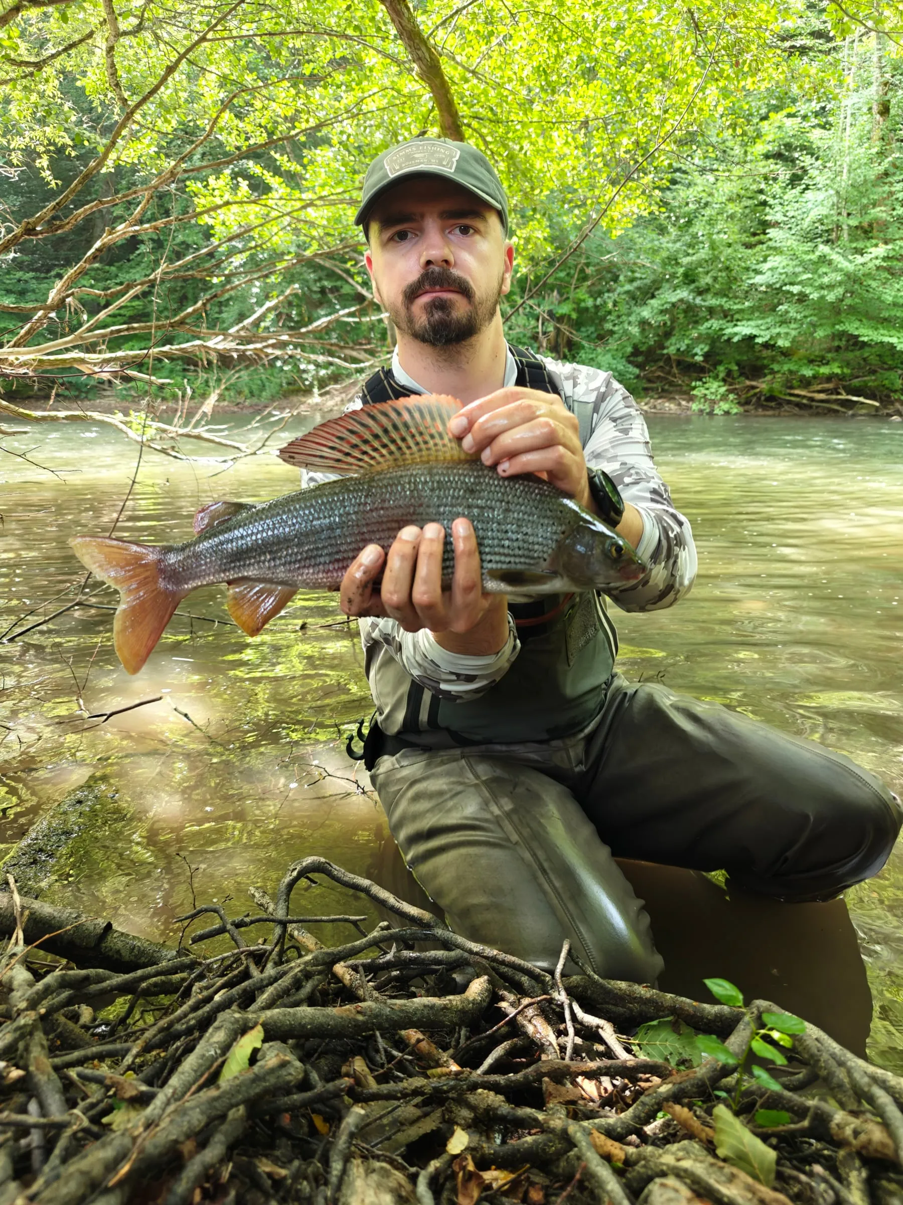 An angler kneeling in the river holding a grayling with its red and grey dorsal fin spread, green trees behind.