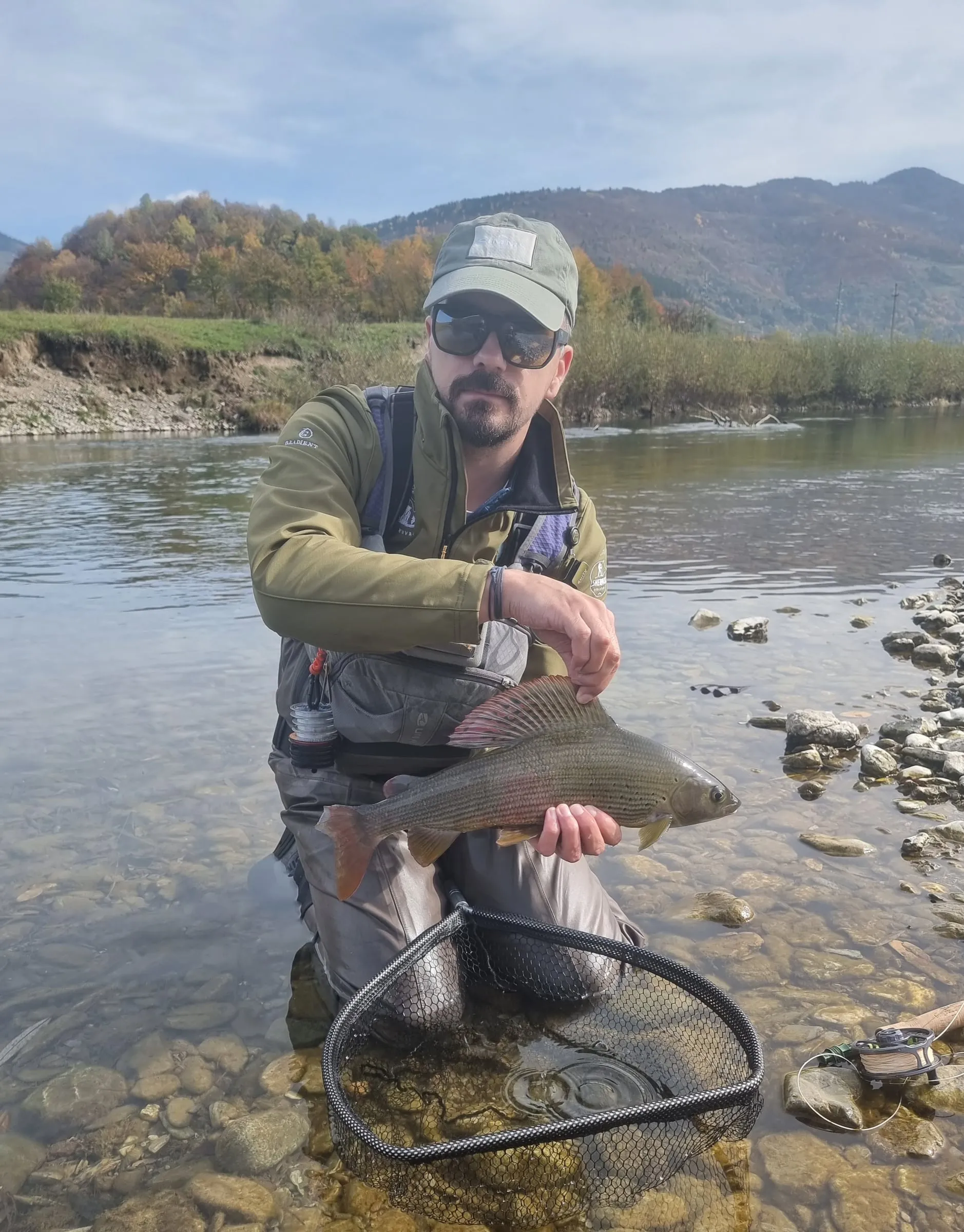 An angler kneeling in clear water holding a grayling over a net, autumn trees and hills behind.