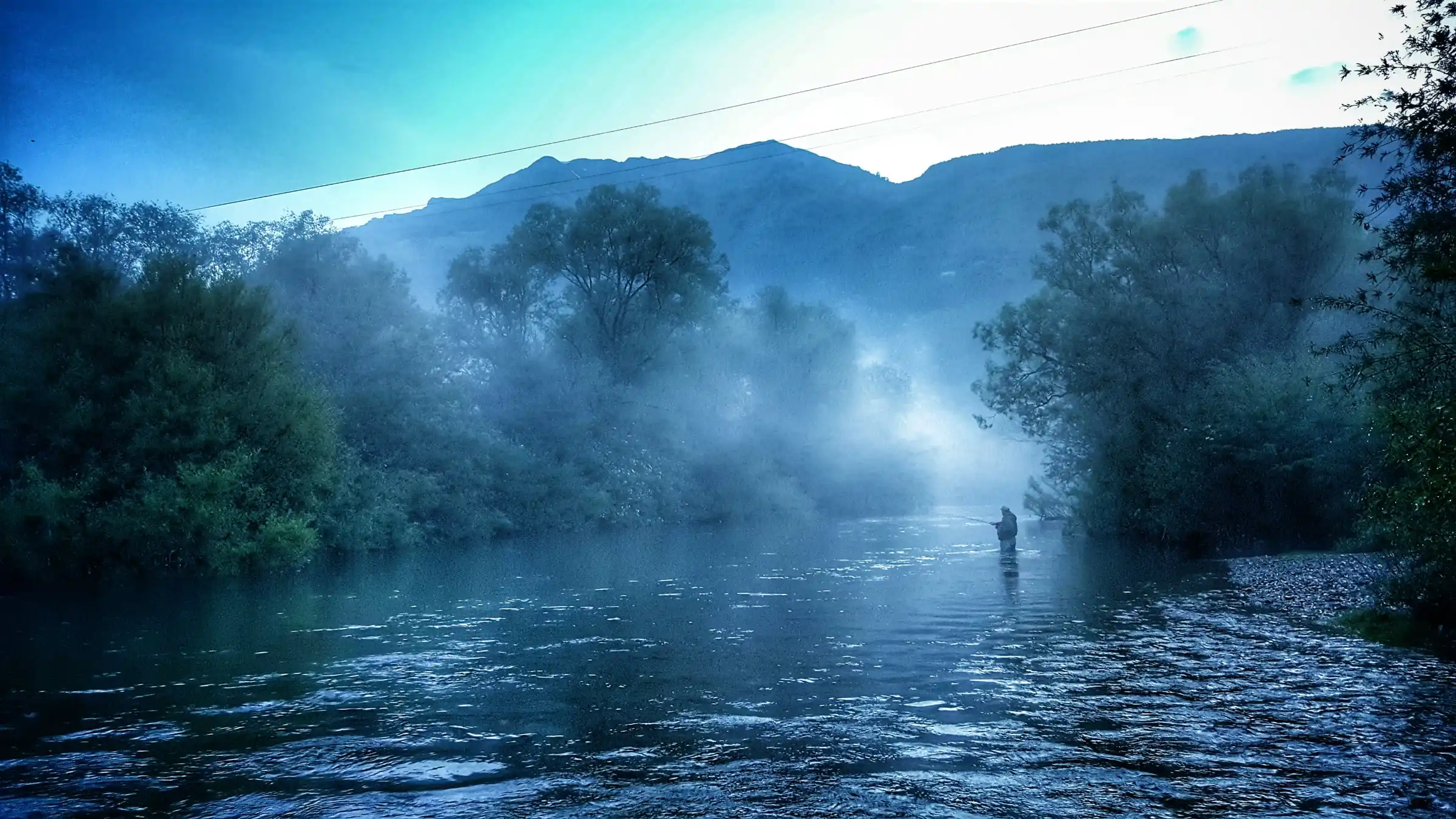 An angler wading the Lim in morning mist between trees, a mountain ridge behind.