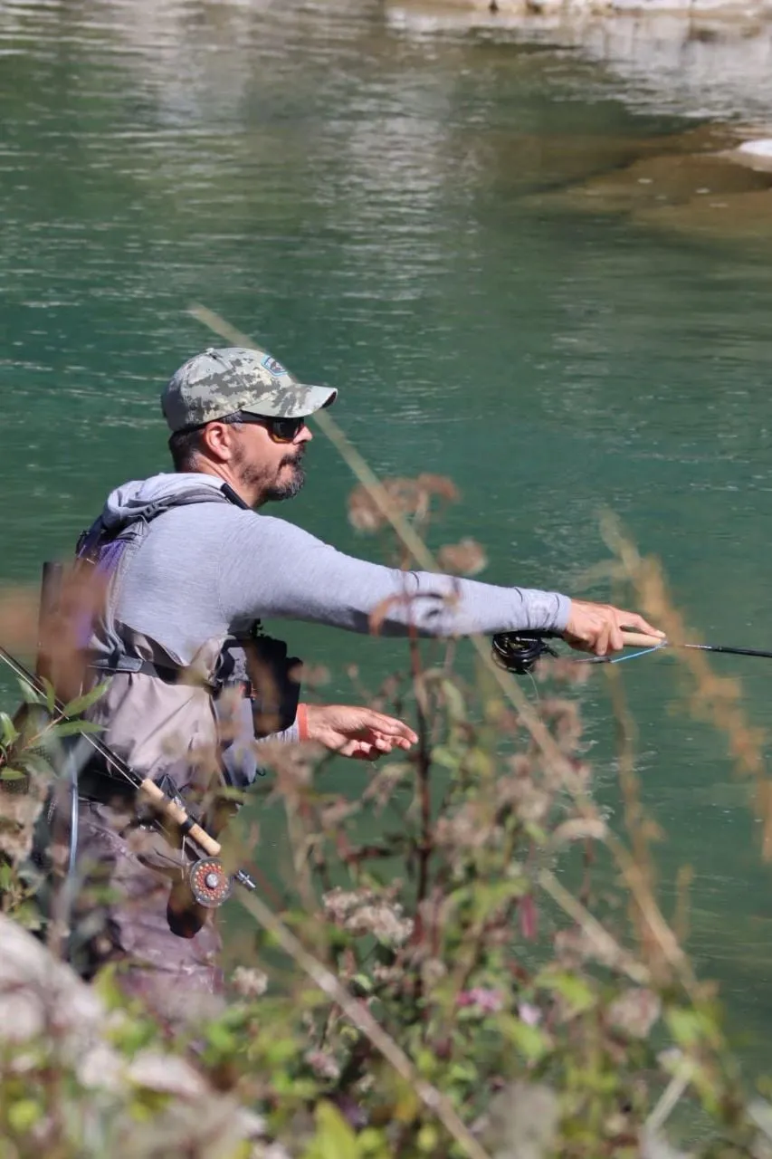 Aleksandar Gasevic casting from the bank of a green river, seen through wild flowers.