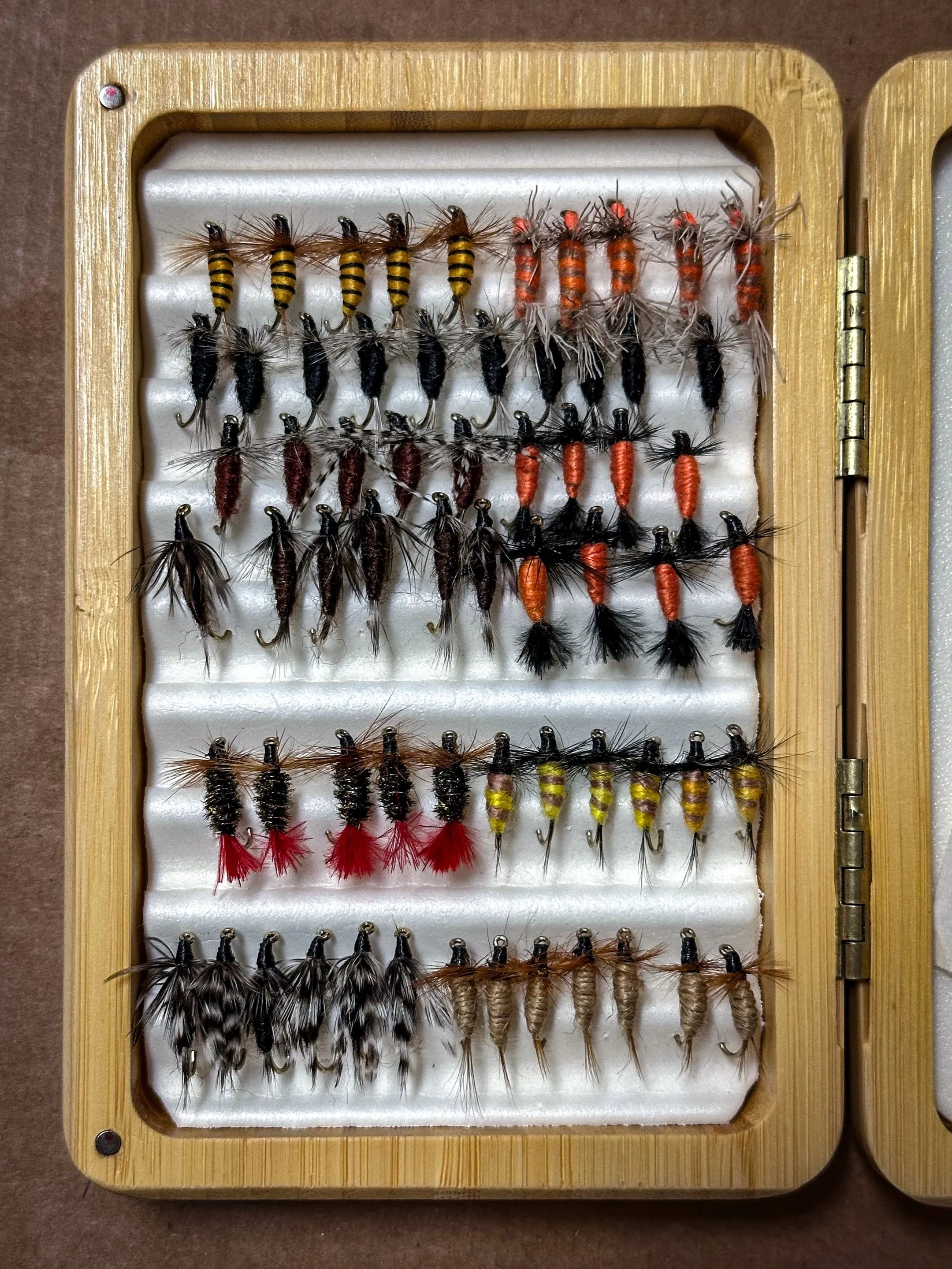 An open bamboo fly box with six rows of small wet flies and nymphs in black, brown, orange and yellow.