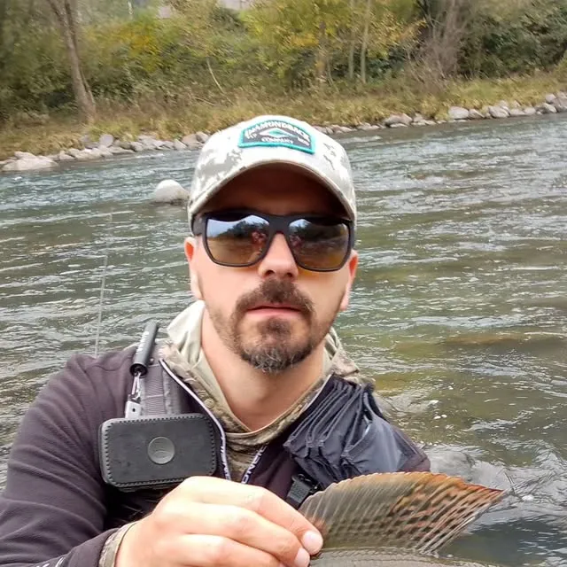 Aleksandar Gasevic standing in a river, holding up a grayling by its dorsal fin.
