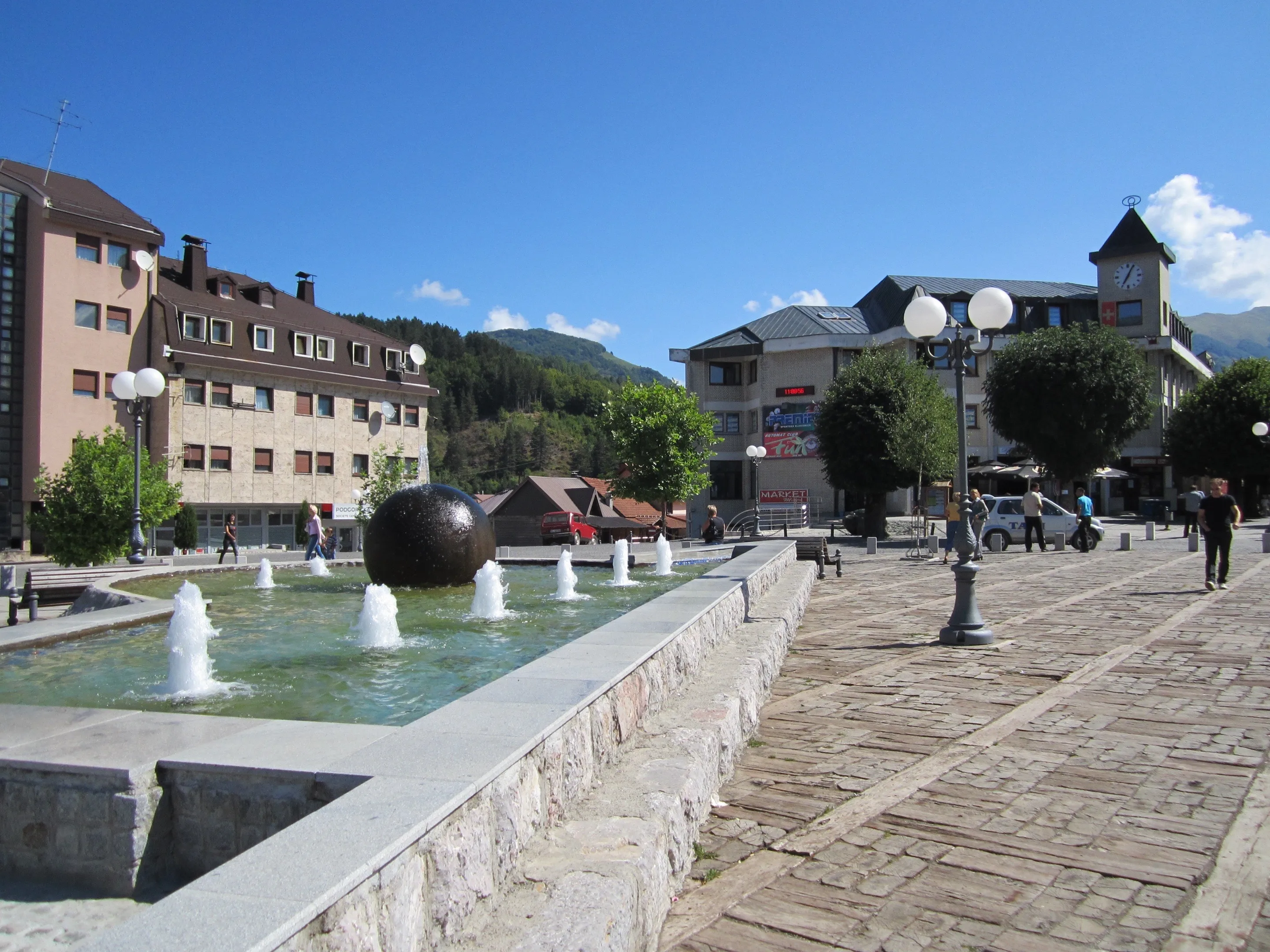 The main square in Kolašin: a fountain with a dark stone sphere, a clock tower and a wooded hill behind the buildings.