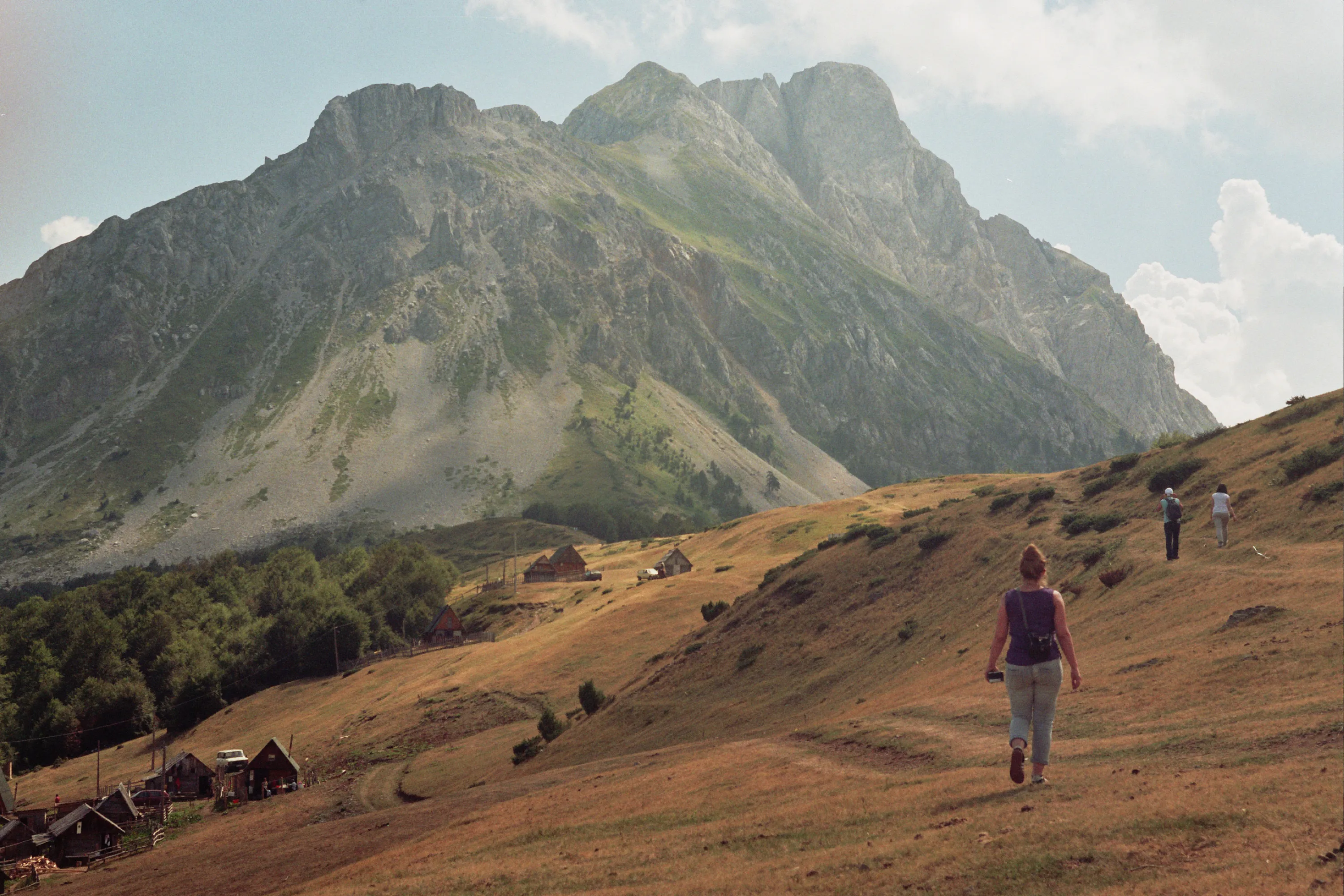 The rock walls of the Komovi above the summer pasture at Štavna, with wooden huts below and walkers crossing dry grass.