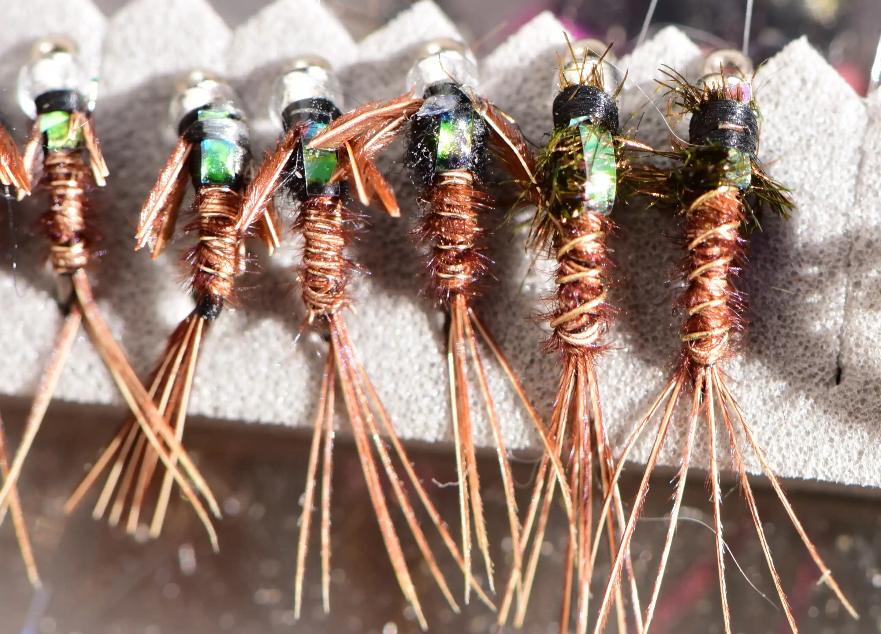 A row of bead-head nymphs with copper-wire bodies and rubber legs standing in a fly box.