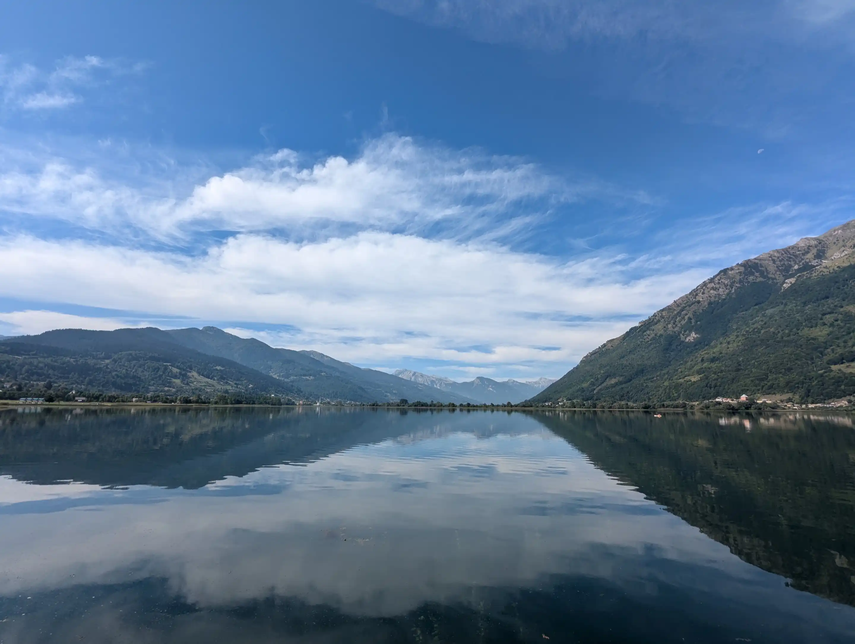 Plav Lake in still weather: flat water mirroring white cloud and blue sky, forested mountain slopes on both sides, and a line of grey rock peaks far off where the valley closes.