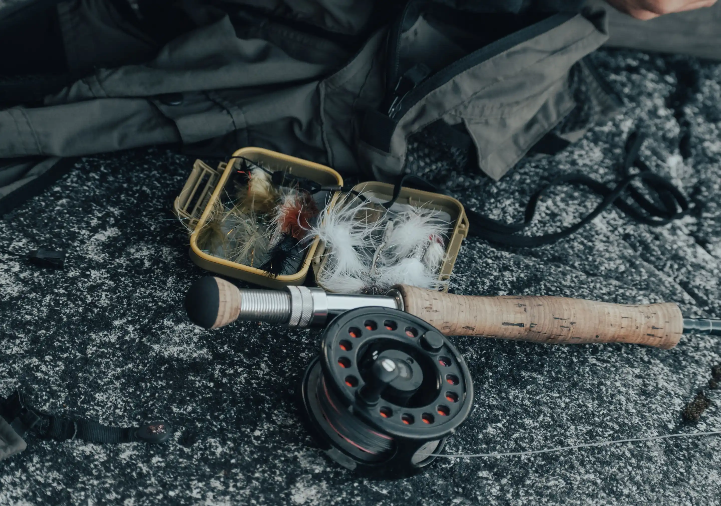 A fly rod, a reel and an open box of flies laid out on dark stone.