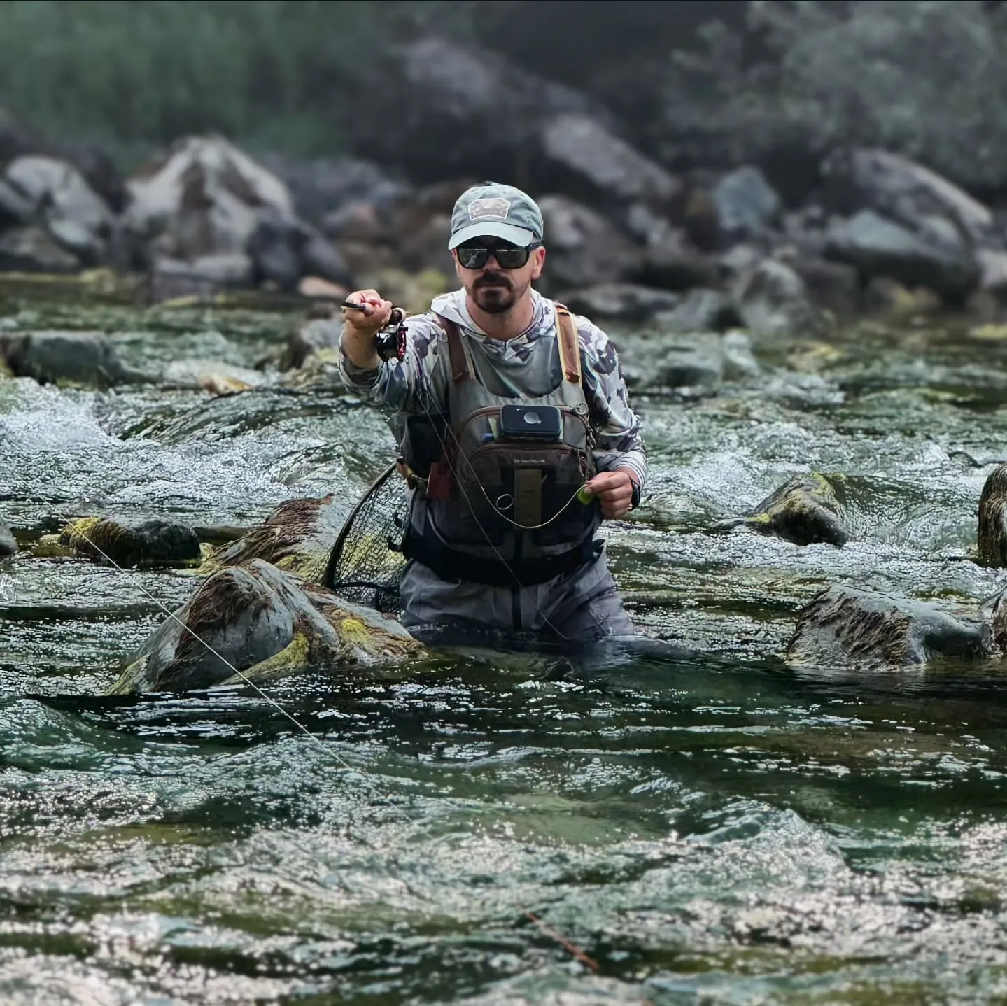 Aleksandar Gasevic wading waist-deep through a fast boulder run, fly rod held out, a landing net at his side.